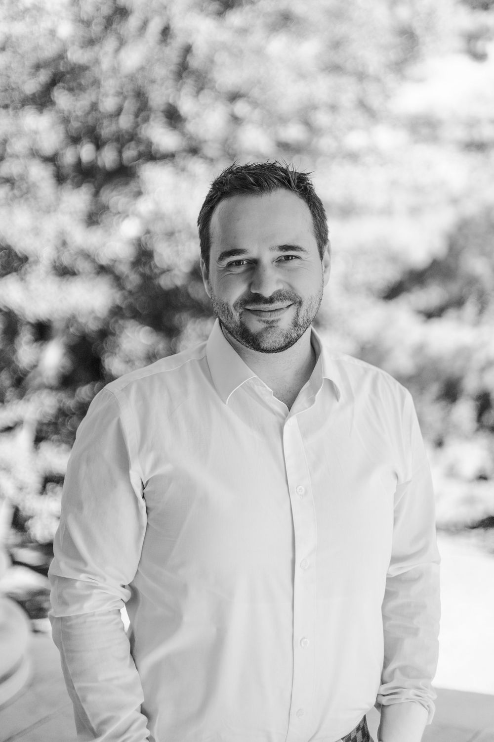 Black and white portrait of a smiling man in a white shirt outdoors with trees in the background.