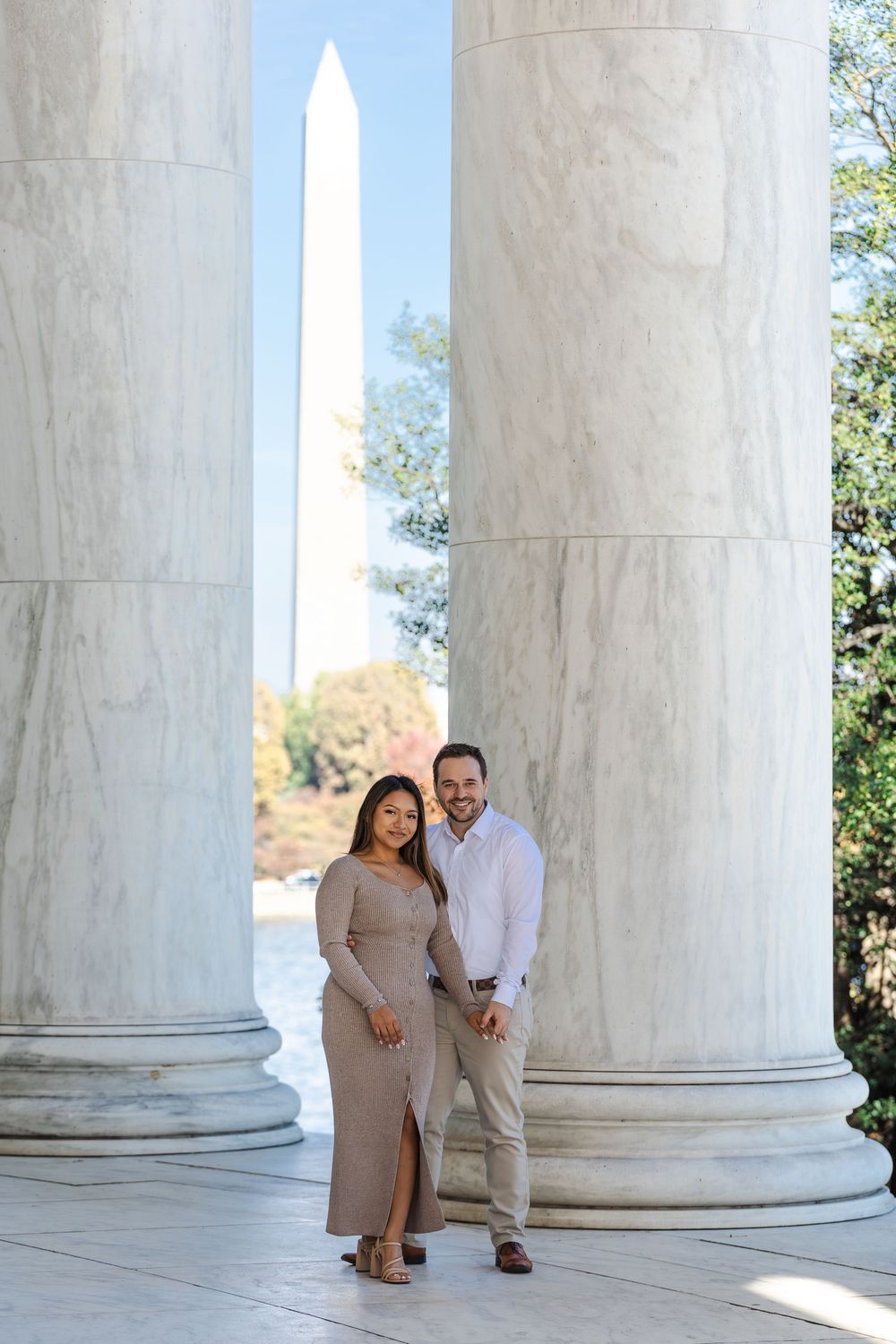 Couple poses between marble columns at Jefferson Memorial with Washington Monument visible in background.