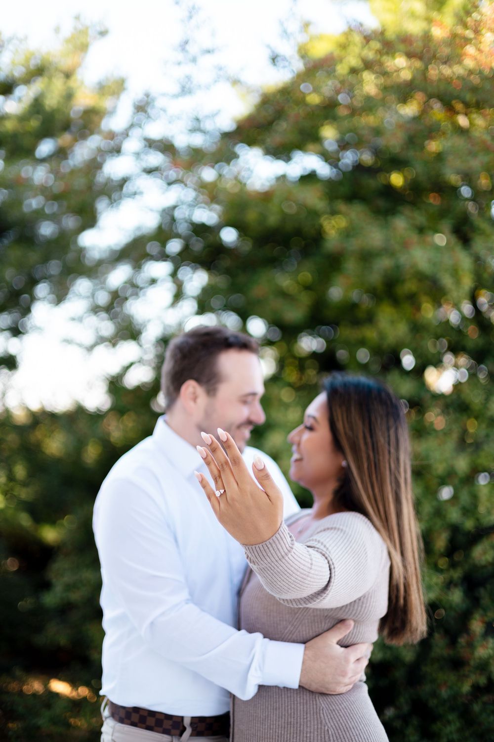 Couple embracing outdoors, woman showing engagement ring, smiling at each other with green trees in background.