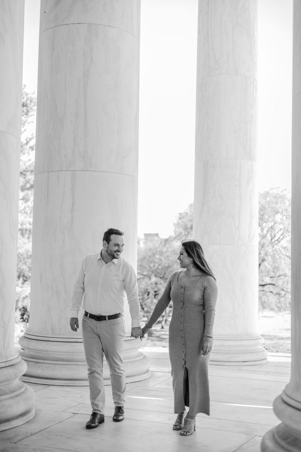 Couple holding hands and smiling between large marble columns at the Jefferson Memorial in black and white.