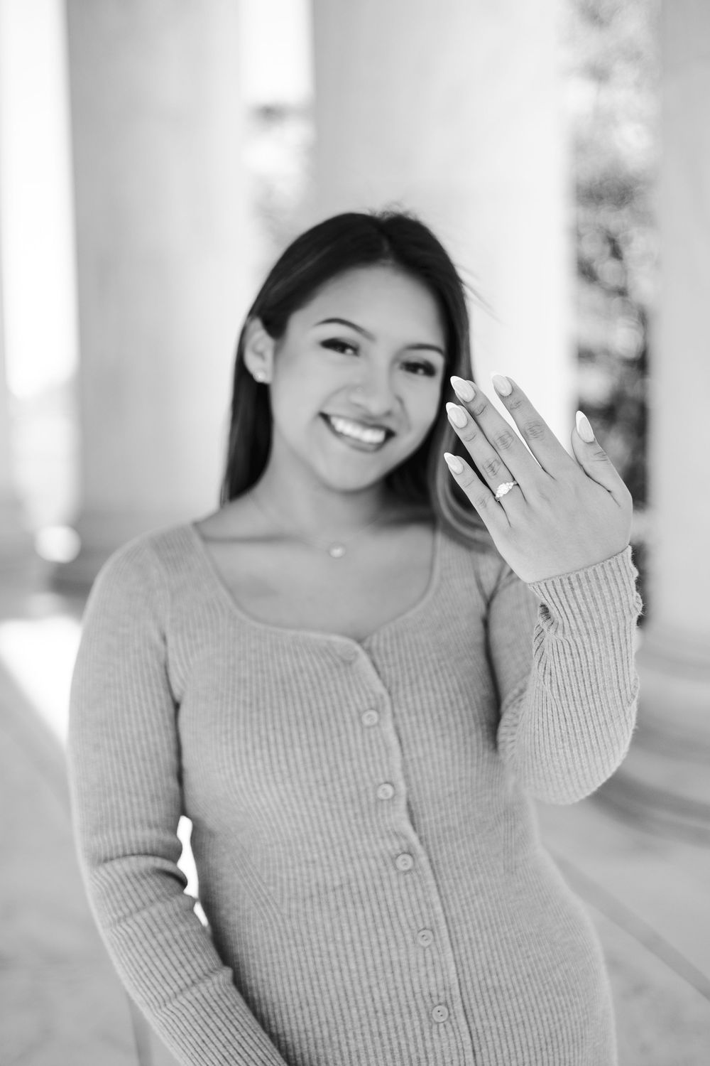 Young woman smiling and showing off an engagement ring on her hand, black and white photo with columns in background.