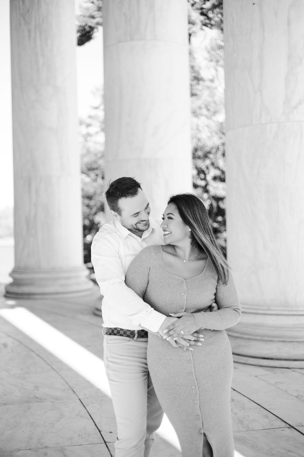 Black and white photo of a smiling couple embracing between large white columns outdoors.