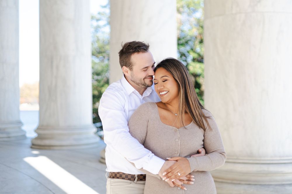 Couple embracing romantically between white columns, woman smiling as man holds her from behind outdoors.