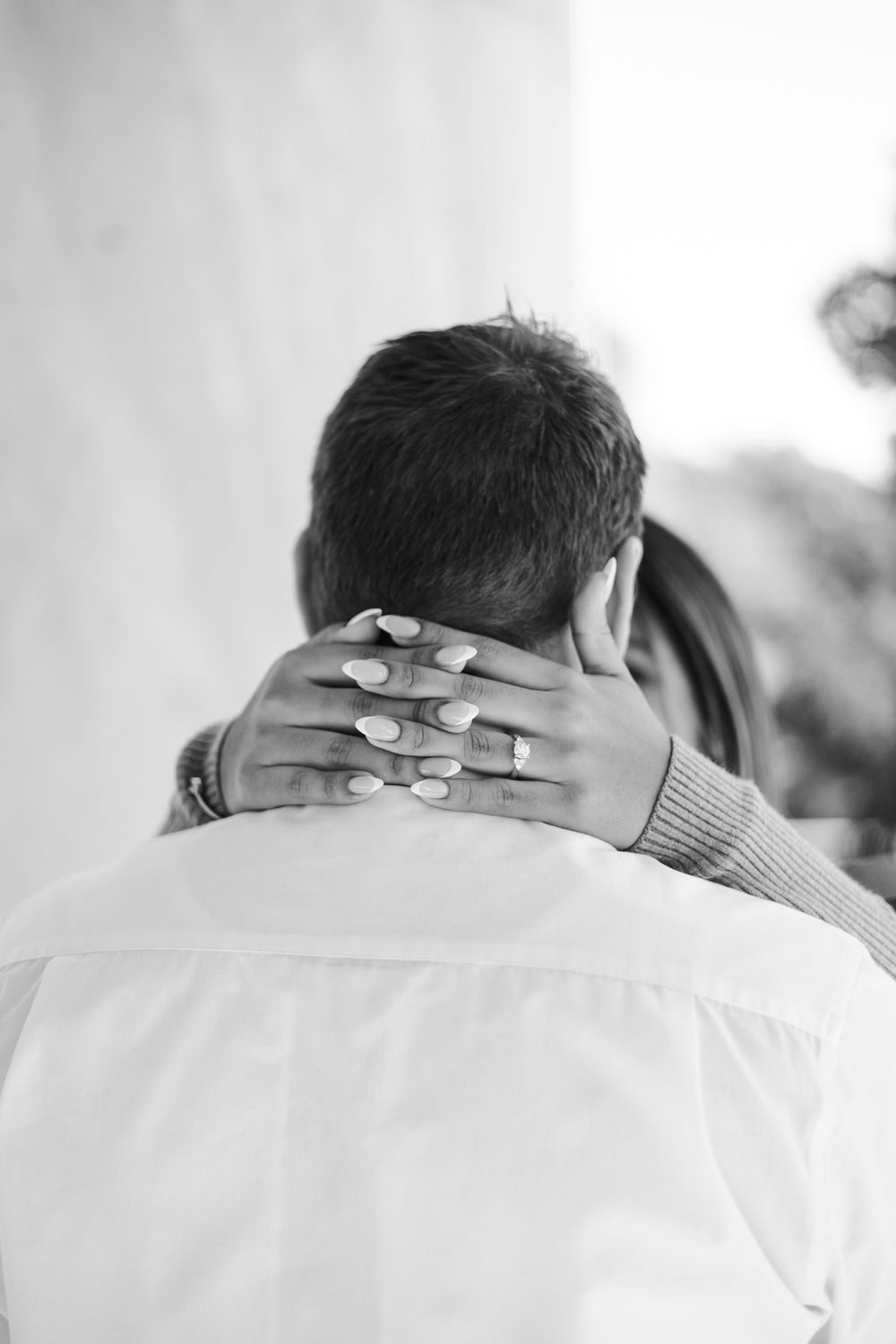 Black and white photo of a woman's hand with engagement ring resting on a man's neck during an embrace.