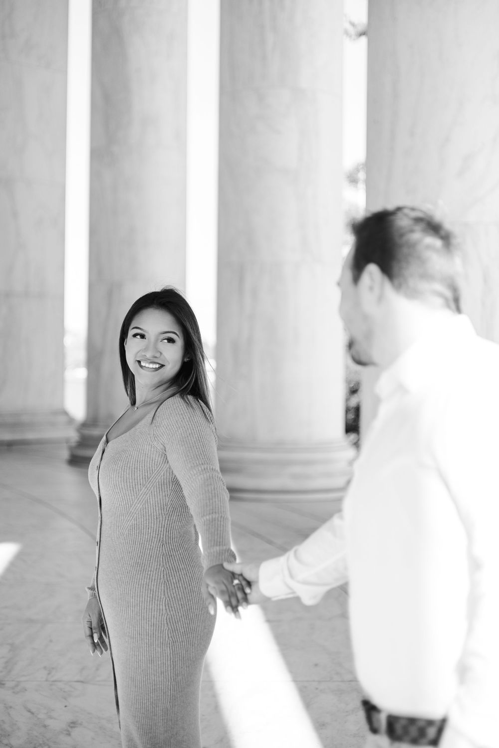 Black and white photo of a smiling woman in a dress holding hands with a man near large marble columns.