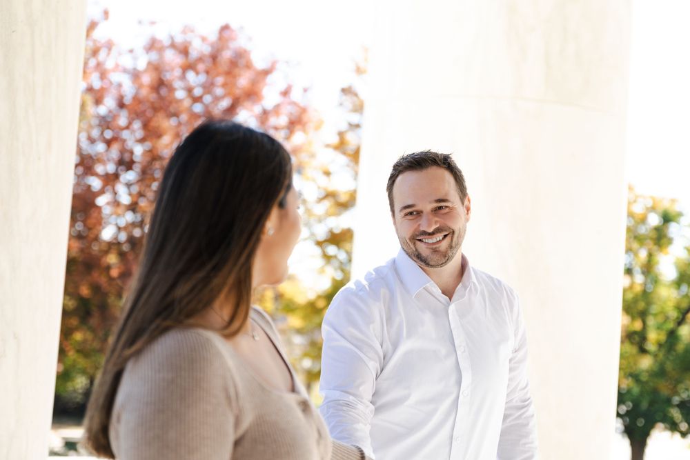 A smiling man in a white shirt shakes hands with a woman outdoors near white columns with autumn trees.