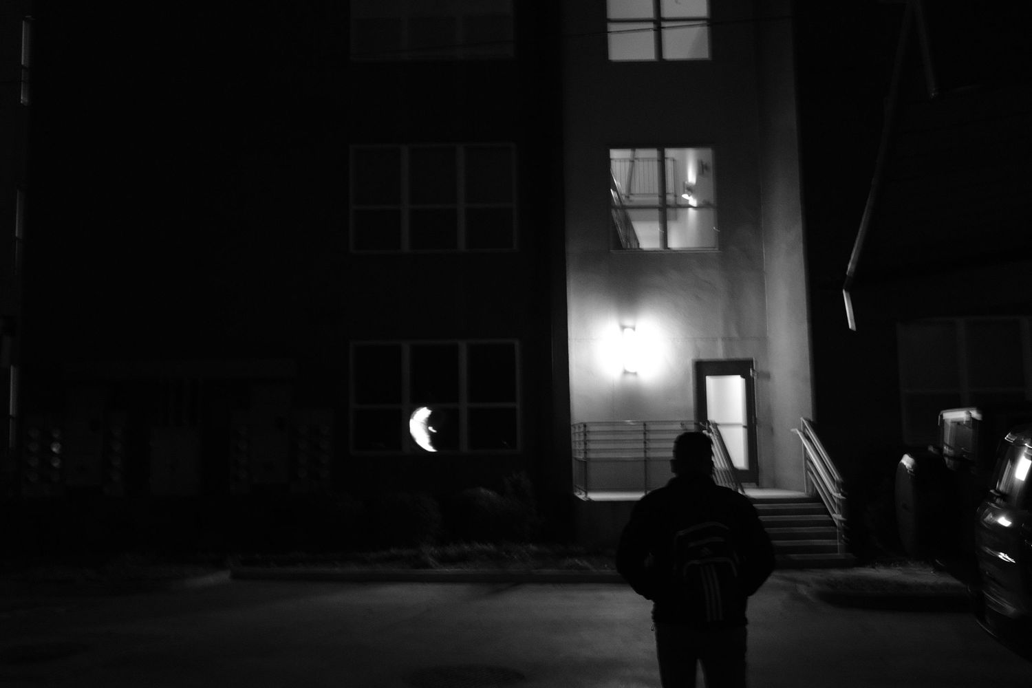 A lone figure stands outside a dark building at night in black and white photography.