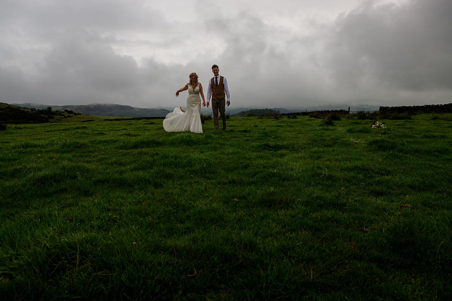 Two figures in wedding attire stand together on a dramatic green hilltop under moody storm clouds in the Lake District