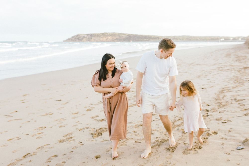 Family of four walking on a sandy beach, parents holding children, ocean waves in the background.