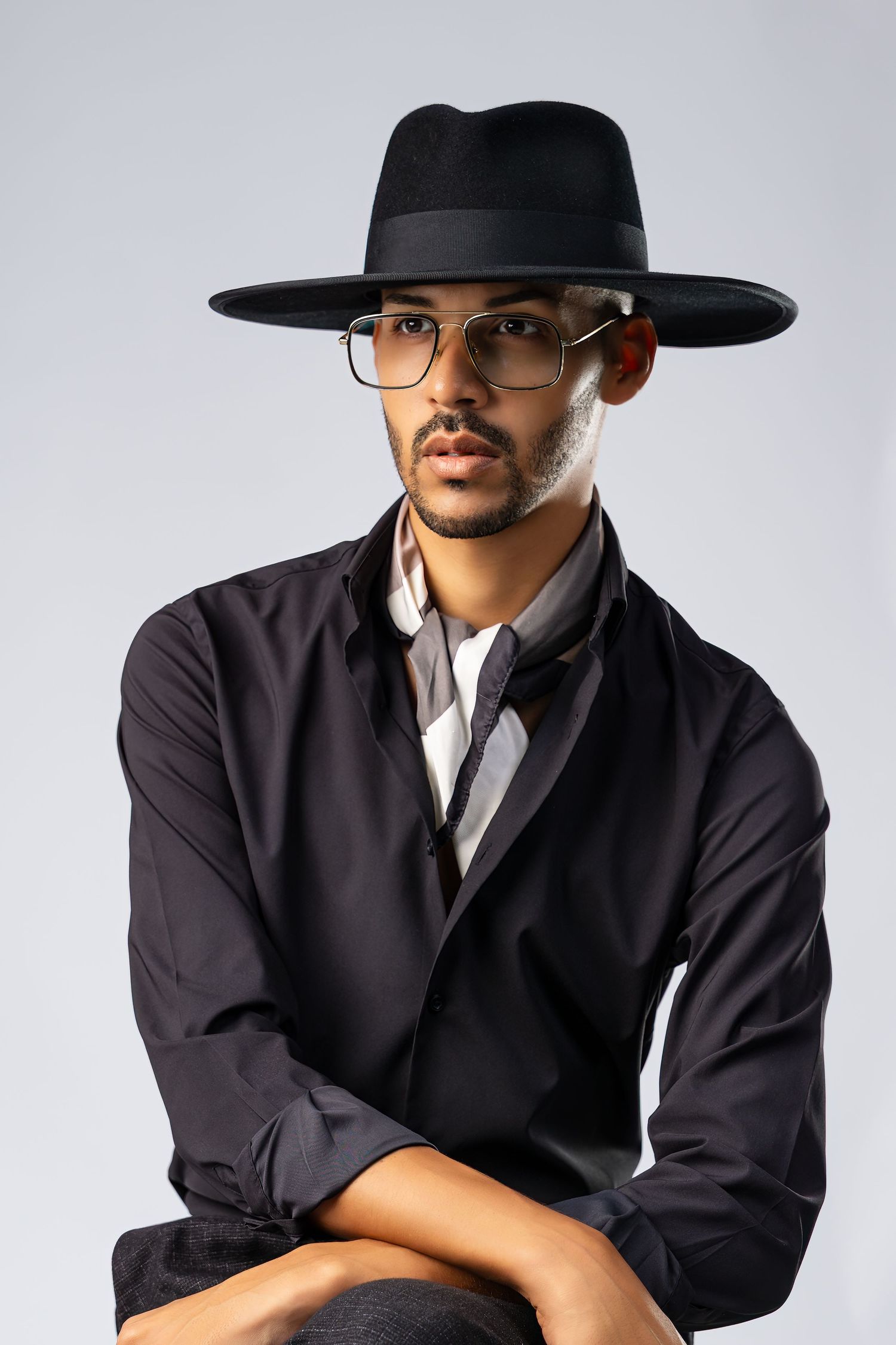 Stylish young man in black hat, glasses, and jacket poses confidently against gray background.
