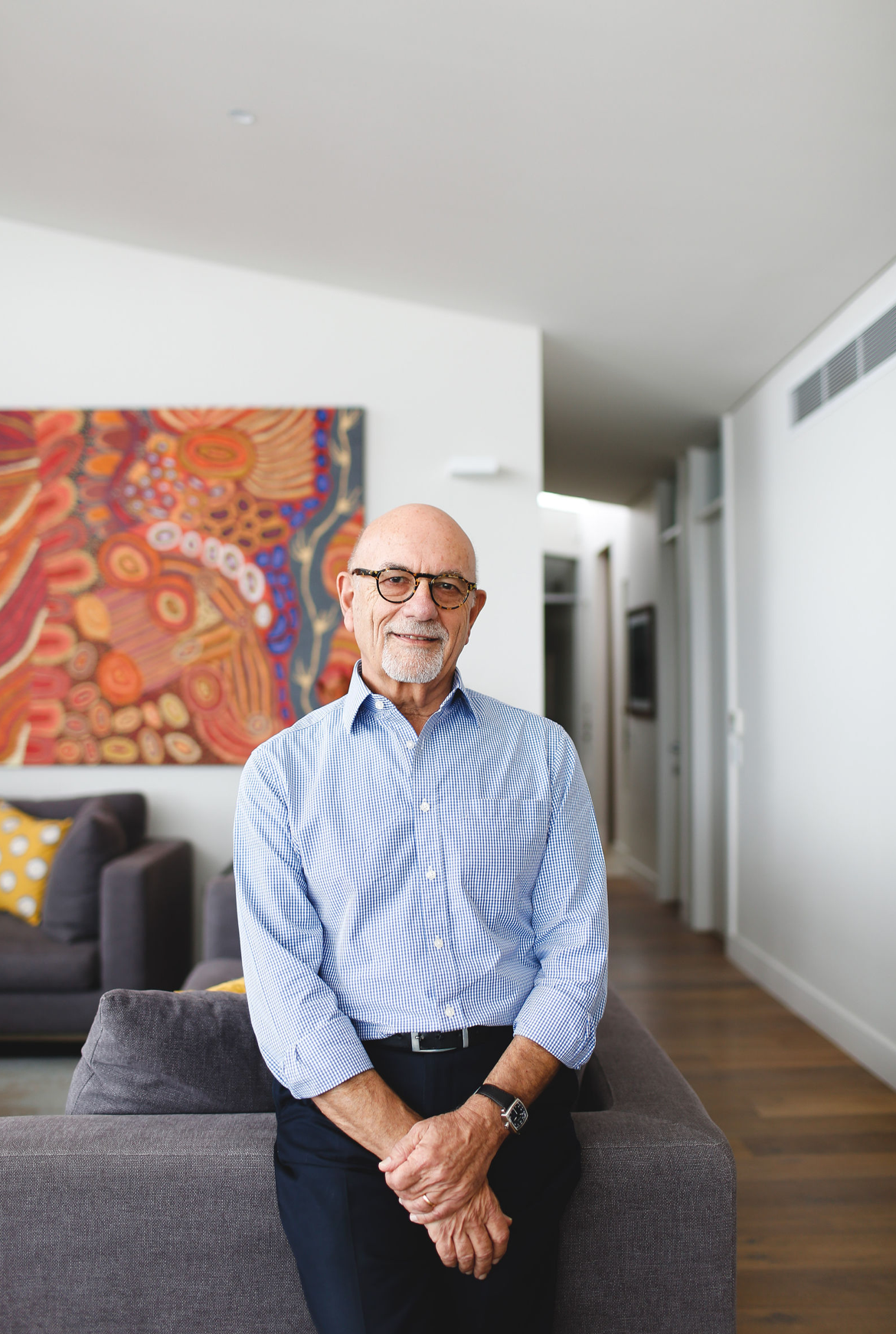 Elderly man with glasses sitting on a gray sofa in a modern home with colorful Aboriginal art on the wall.