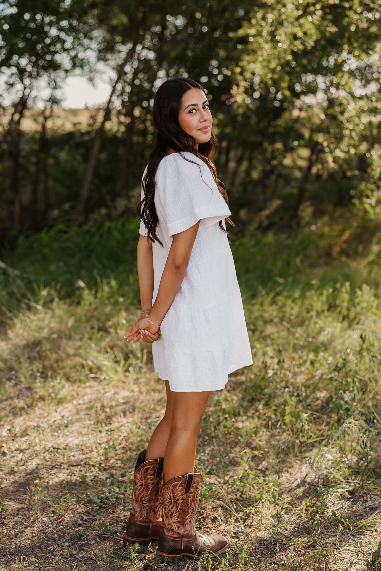 A senior girl poses with arms behind back wearing white dress and cowboy boots in Tulia Texas for senior photos