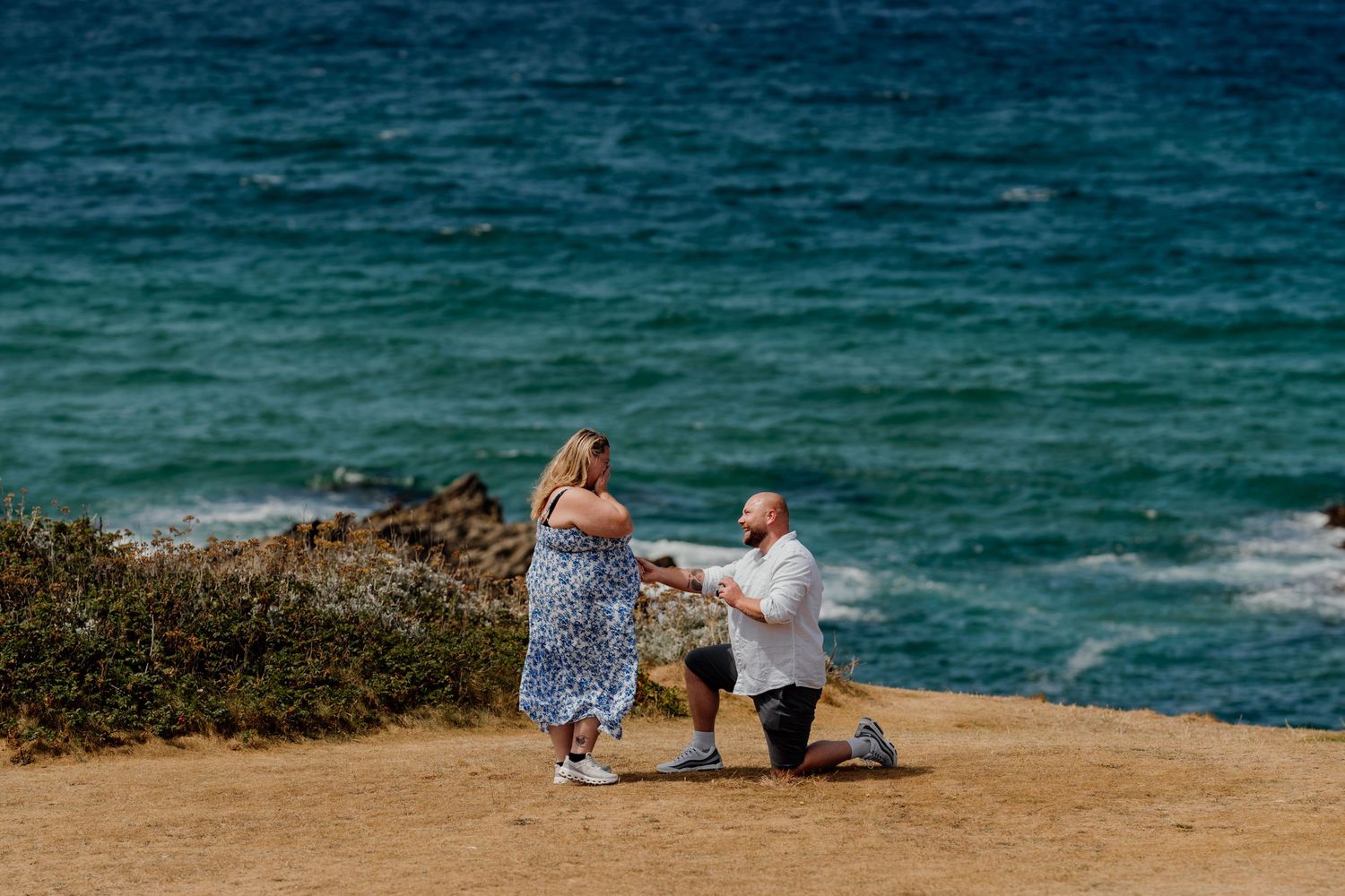 Man proposes on one knee on the cliffs overlooking the headland hotel in Newquay with little fistral behind them.