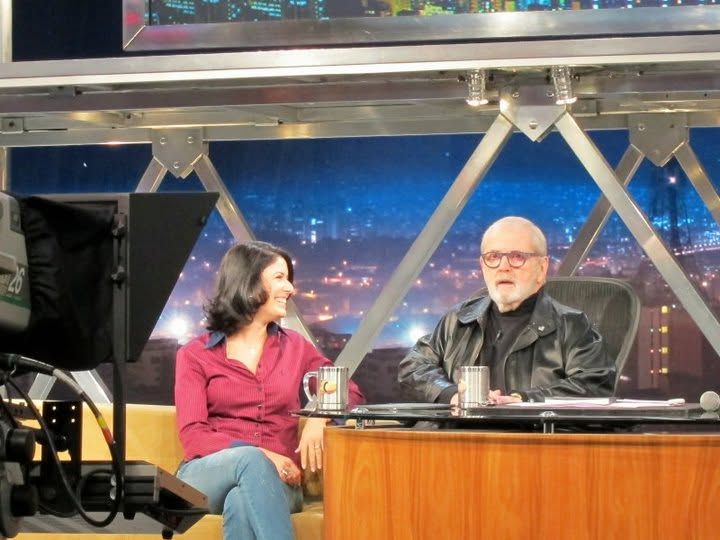Two people conversing on a late-night TV talk show set with city skyline backdrop and wooden desk.