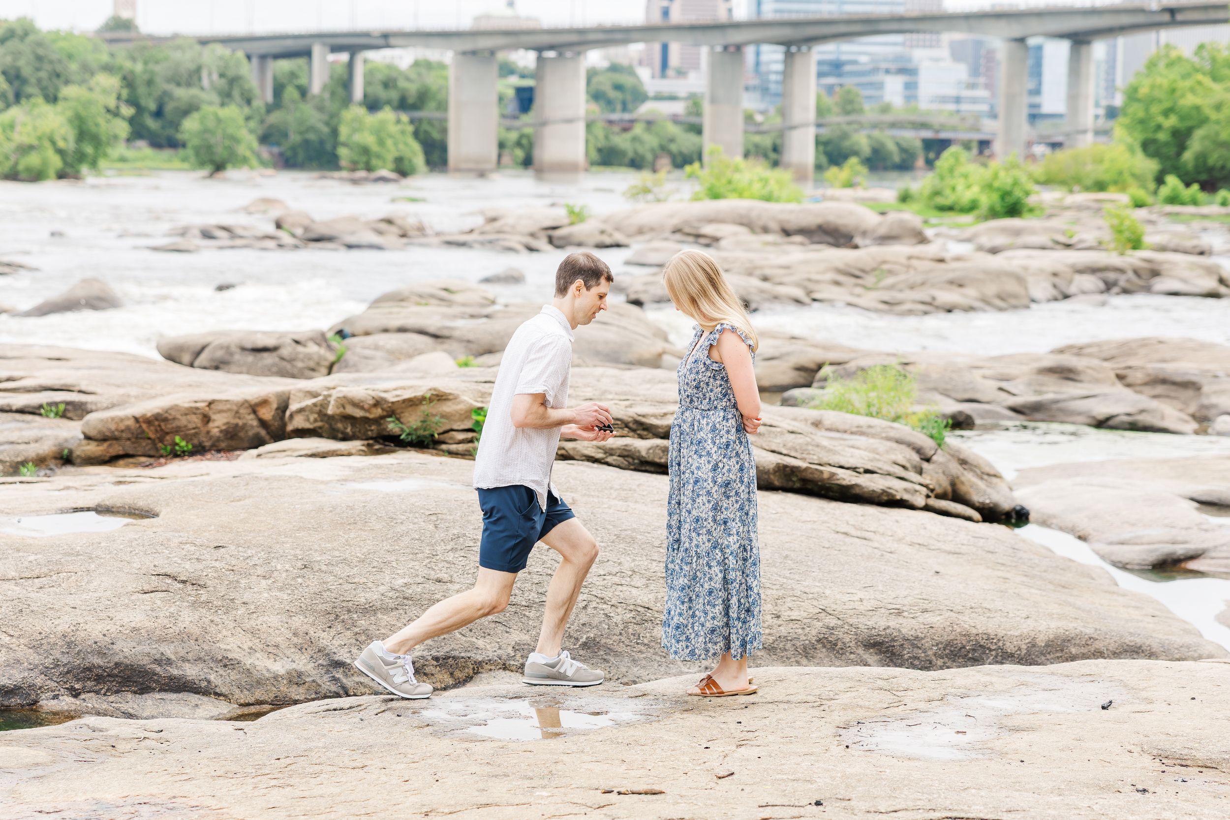 Couple walking hand in hand on rocky riverbank with bridge and city skyline in background.