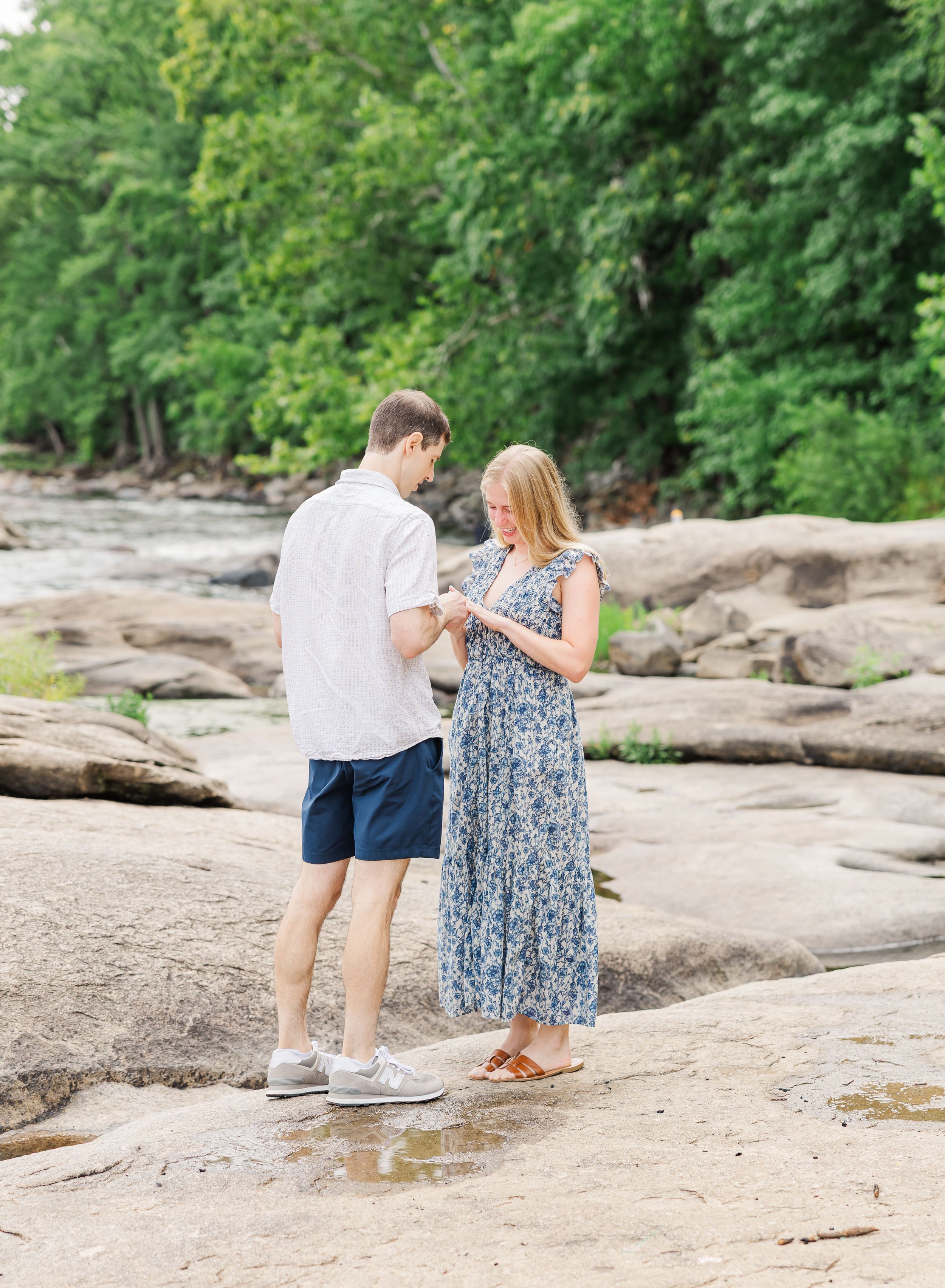 Couple standing on rocky riverbank surrounded by lush green trees, man in white shirt and navy shorts with woman in floral dress.
