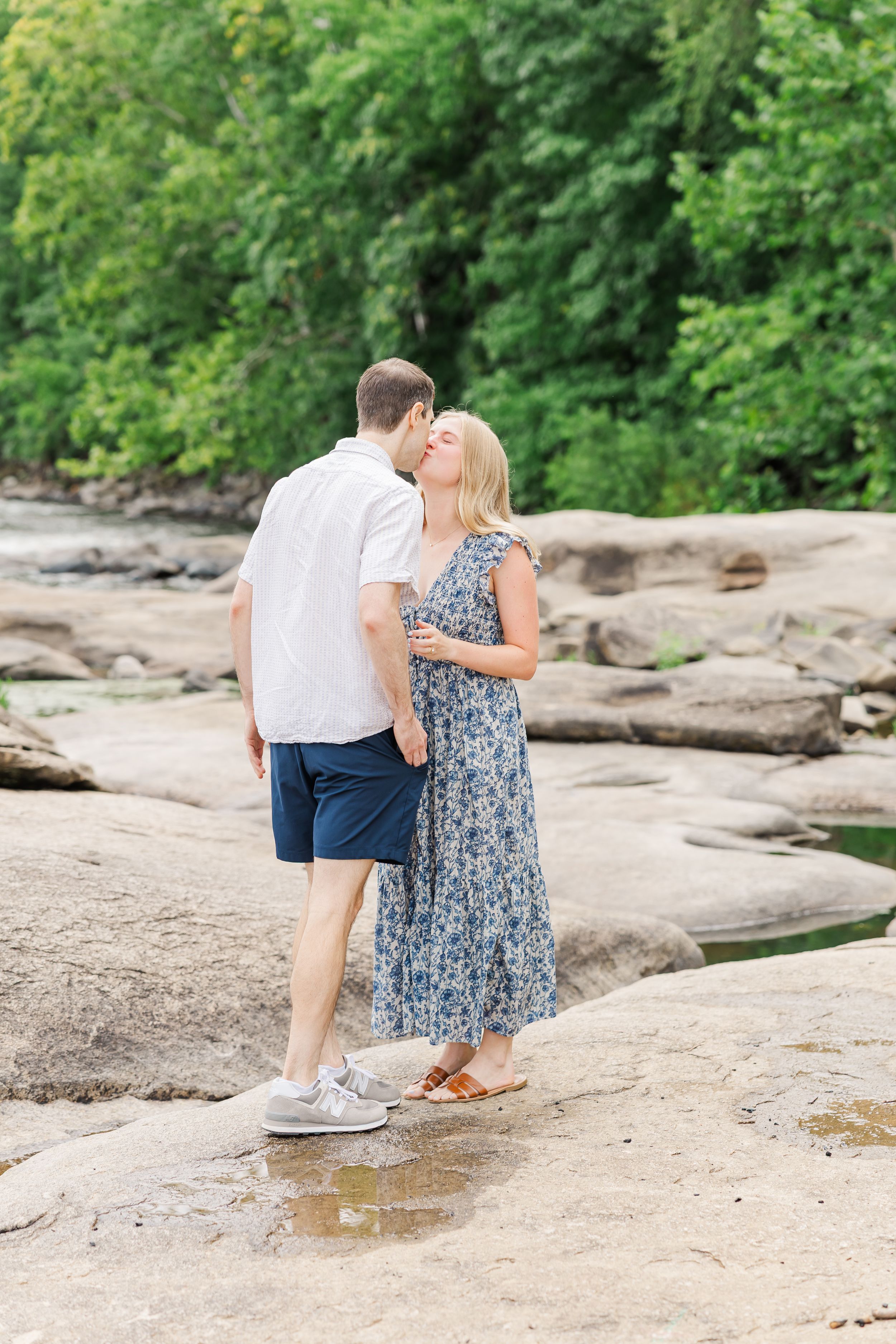 Couple sharing a romantic kiss on rocky terrain outdoors, woman in floral maxi dress, man in white shirt and navy shorts.