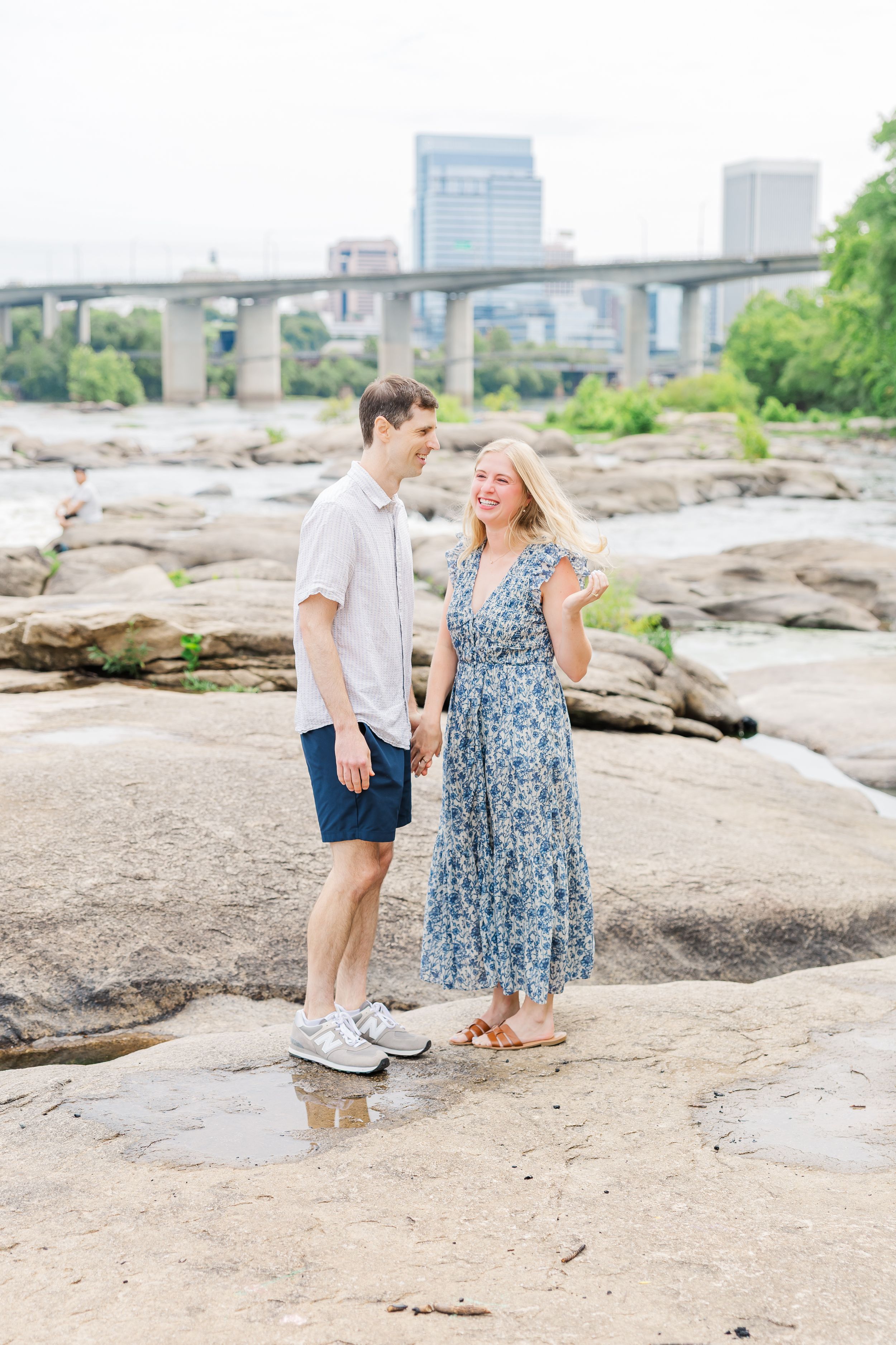Couple holding hands on rocky riverbank with city skyline and bridge in background on a sunny day.