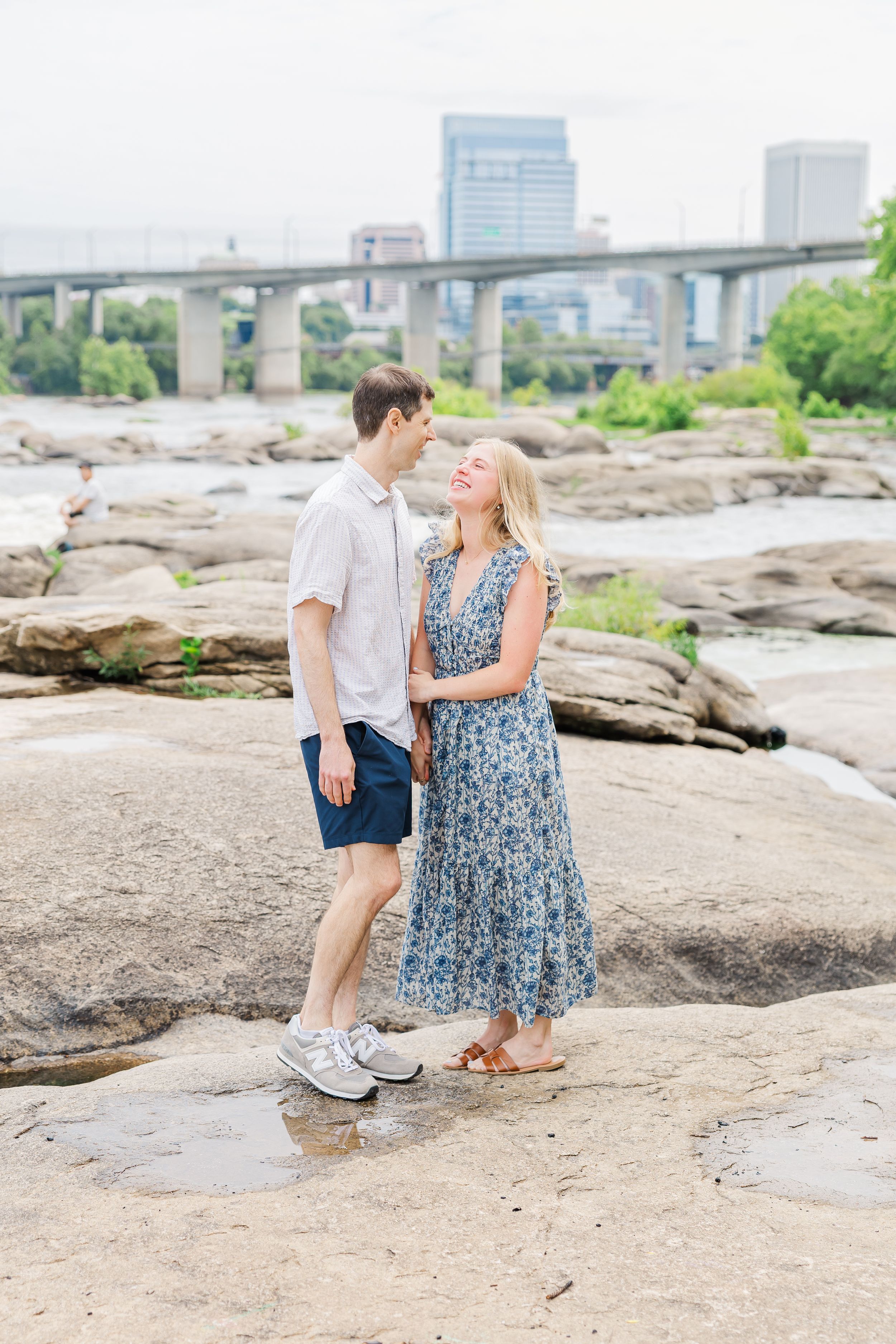 Couple standing on rocks by a river with a city bridge and skyline in the background.