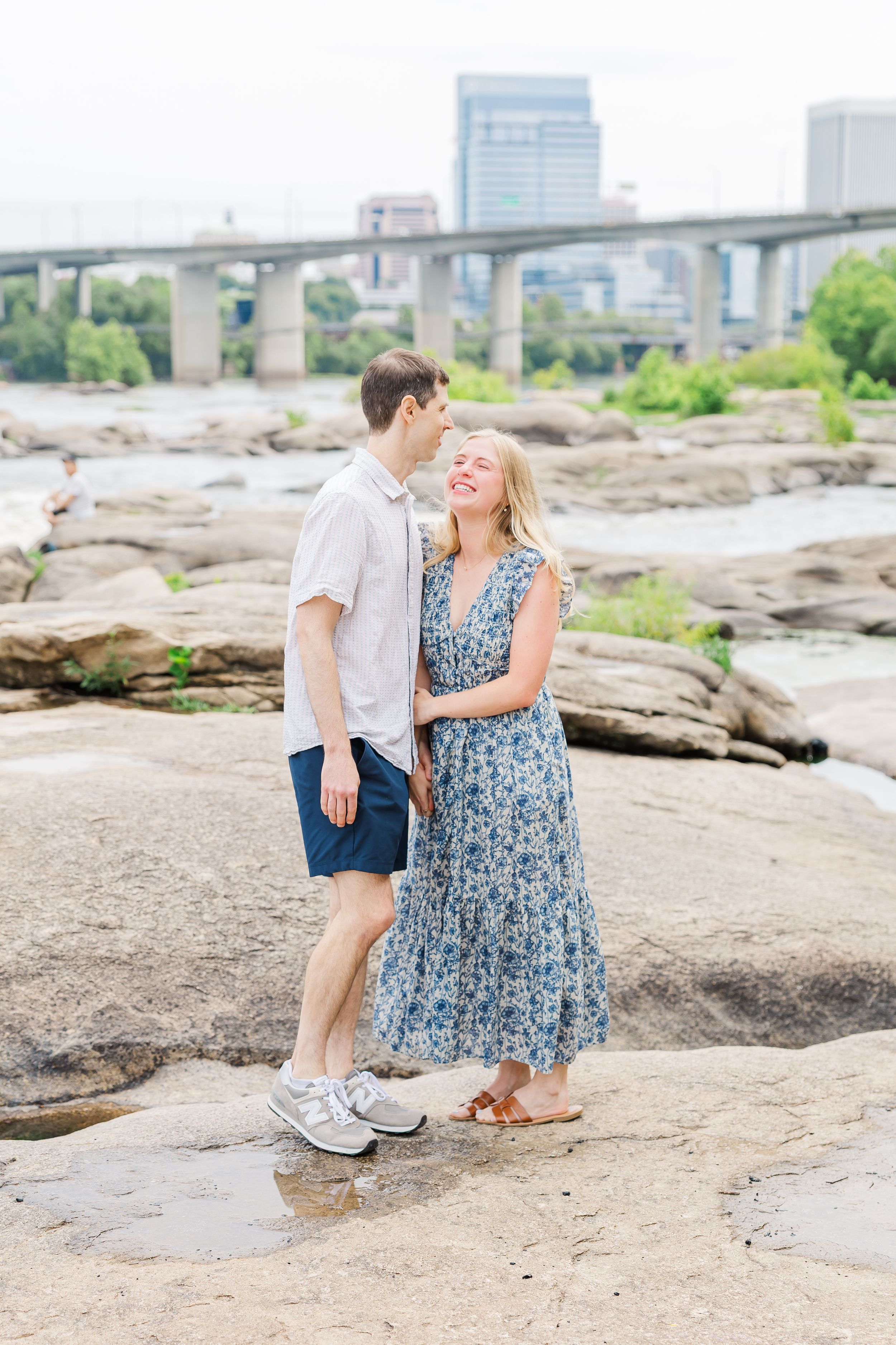 Couple standing on rocks by a river with a city bridge backdrop, woman in floral dress laughing up at man.