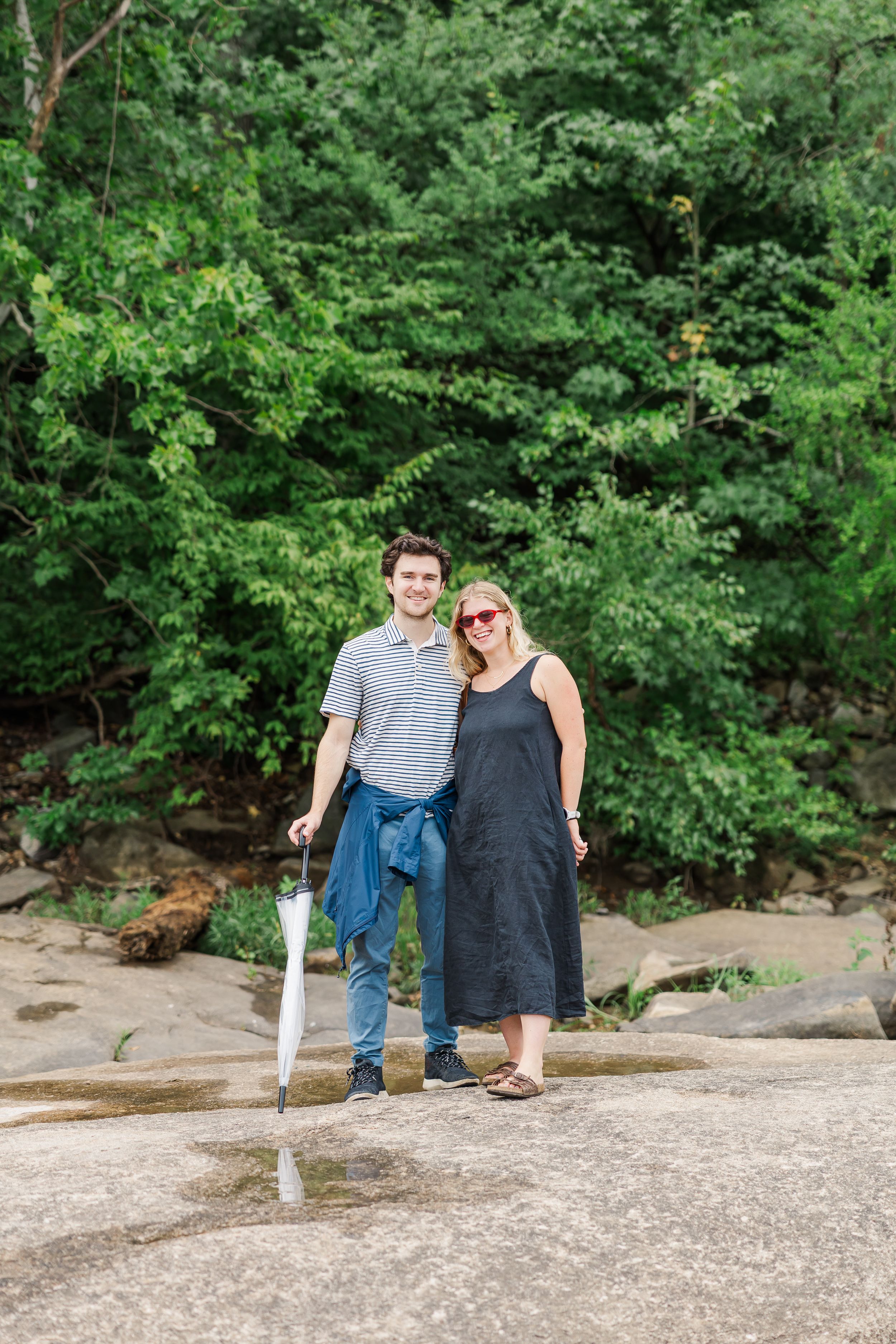 A couple smiles together on rocky terrain, surrounded by lush green trees, man holding umbrella and denim jacket.