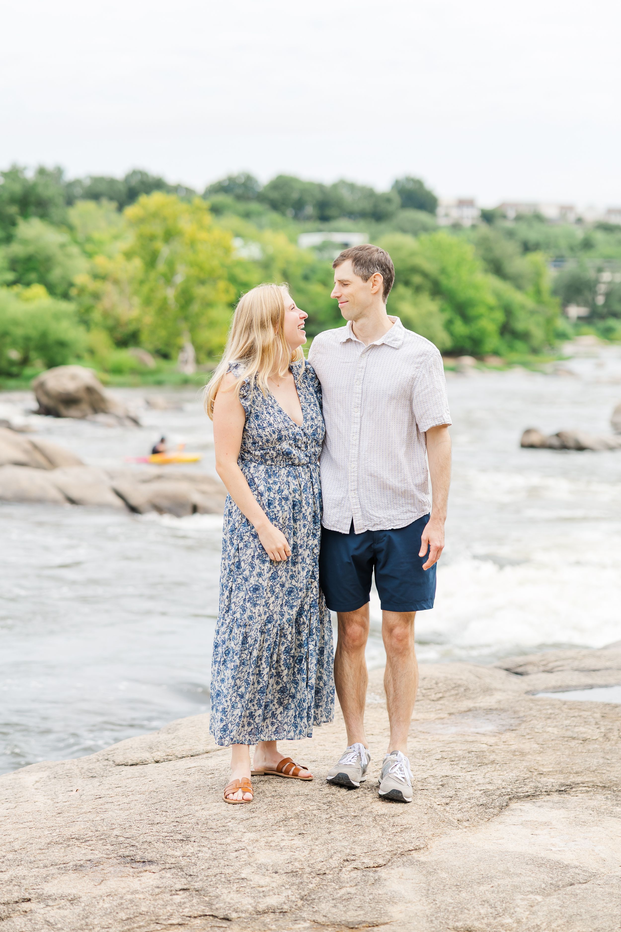 Couple gazing at each other on rocky riverbank, woman in blue floral maxi dress, man in navy shorts.