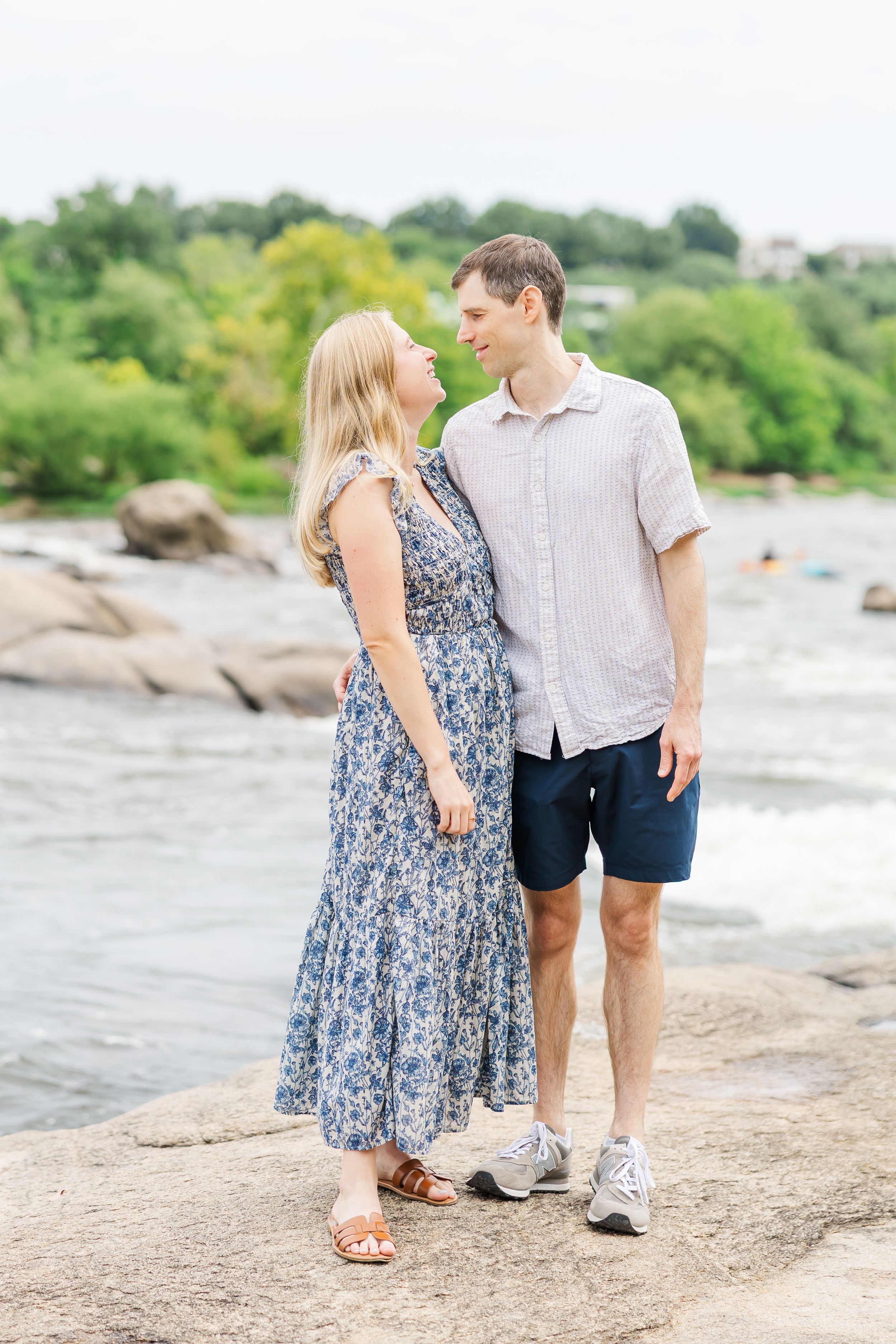 Couple smiling at each other on rocky riverbank, woman in floral dress, man in linen shirt and navy shorts.