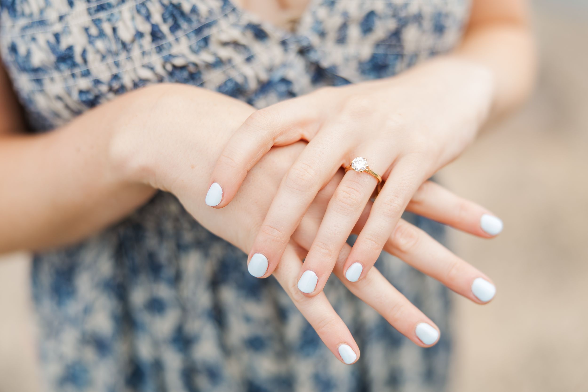 Close-up of woman's hands with light blue manicure and gold ring, wearing a blue floral dress.