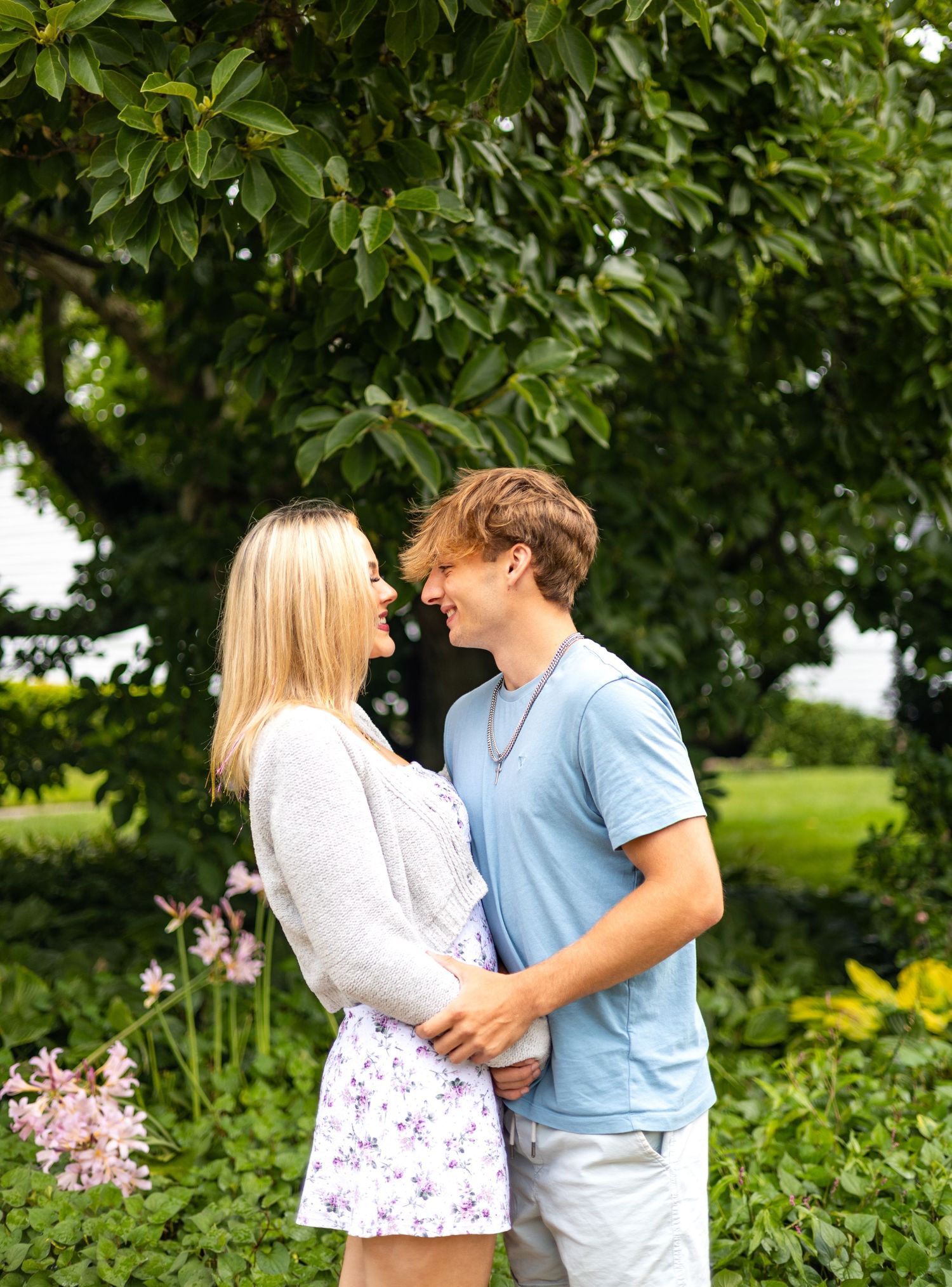 Young couple smiling at each other while embracing under a large green tree.
