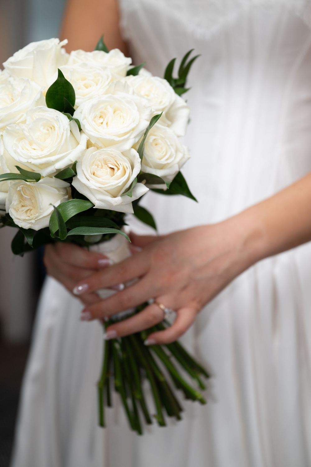 Bride in white dress holding elegant bouquet of white roses with green stems, wearing diamond engagement ring.