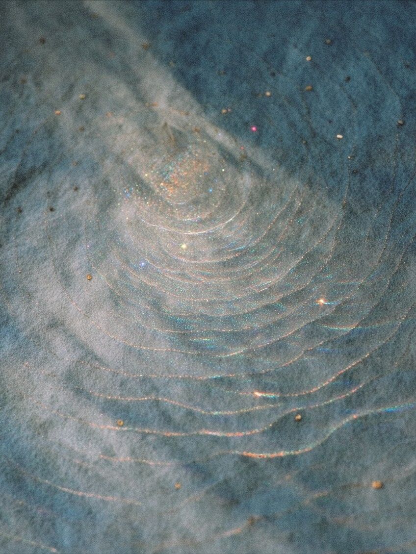 Close-up of swirling concentric rings on a blue textured surface with iridescent glitter particles scattered throughout.