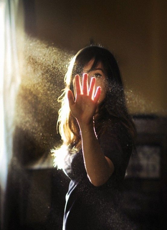 Woman holding glowing hand up to sunlight with golden dust particles floating in a dark moody room.