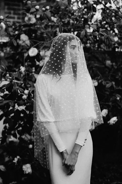 Bride in polka dot veil and white dress stands before blooming flowers in black and white photo.