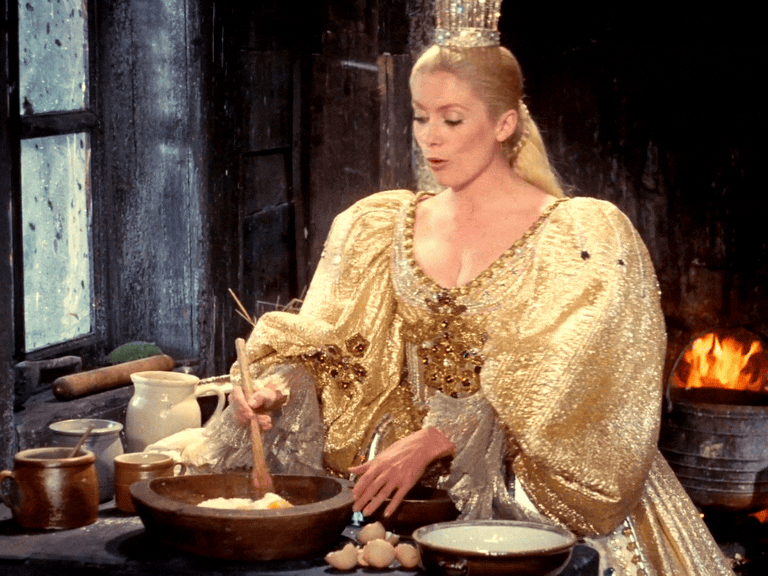 Woman in ornate golden gown cooking in rustic kitchen with fireplace, resembling a fairy tale queen.