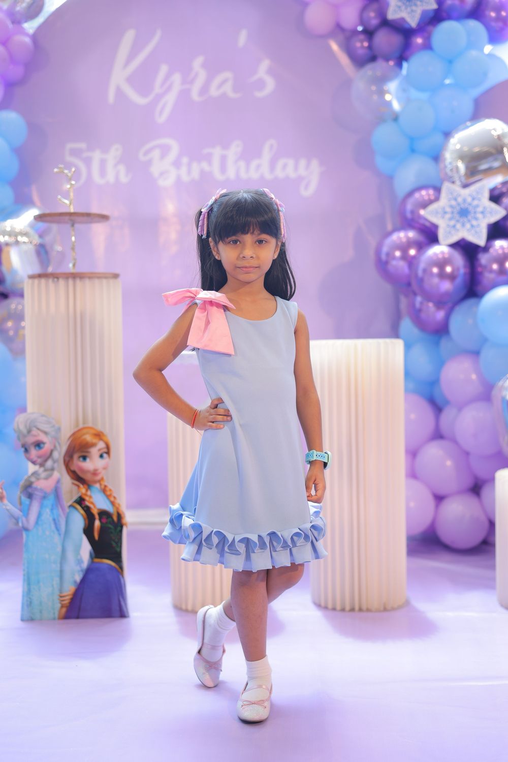Young girl in blue dress poses at Kyra's Frozen-themed 5th birthday party with purple balloon decorations.