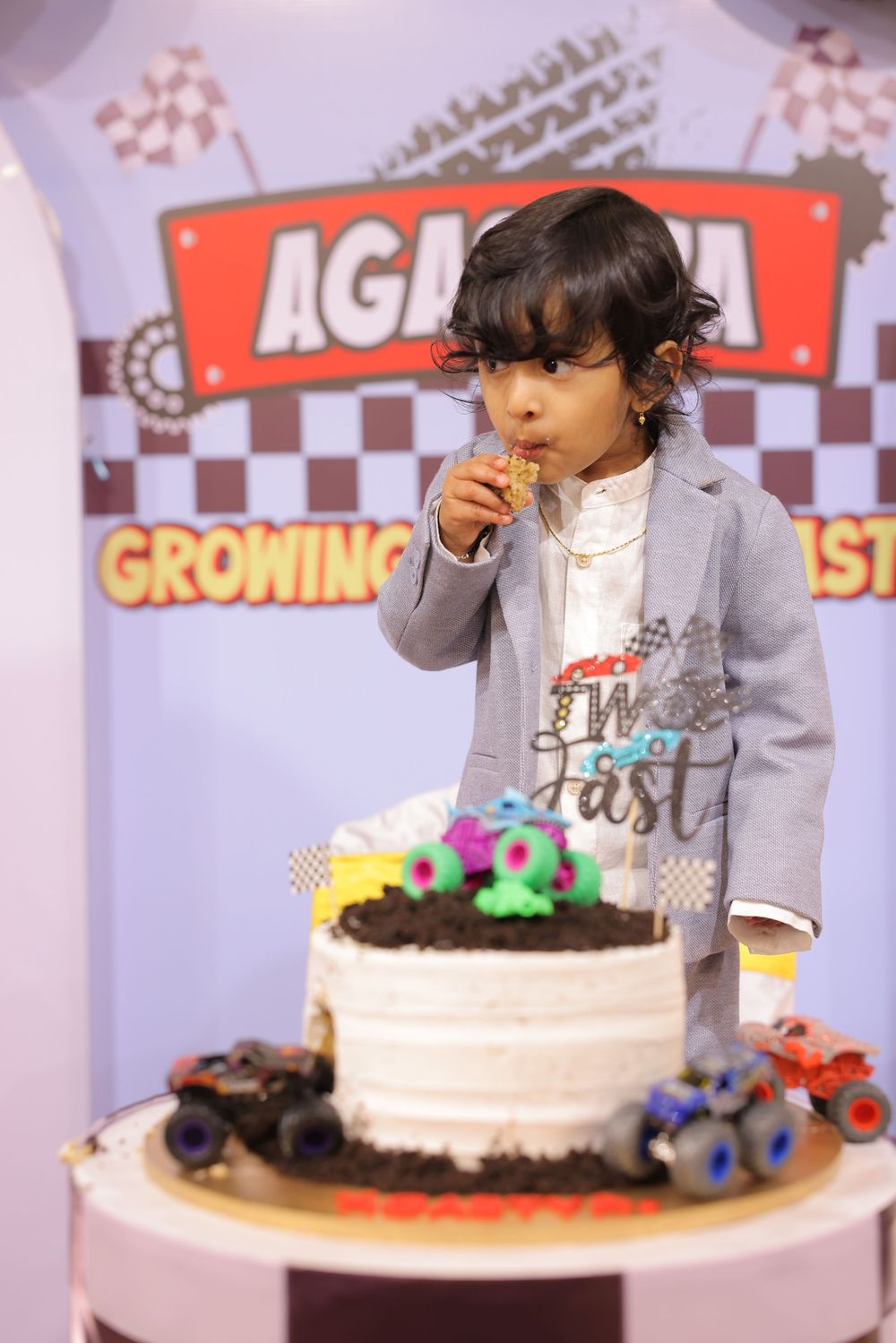 Young boy in gray blazer eating cookie at monster truck themed birthday party with decorated cake.