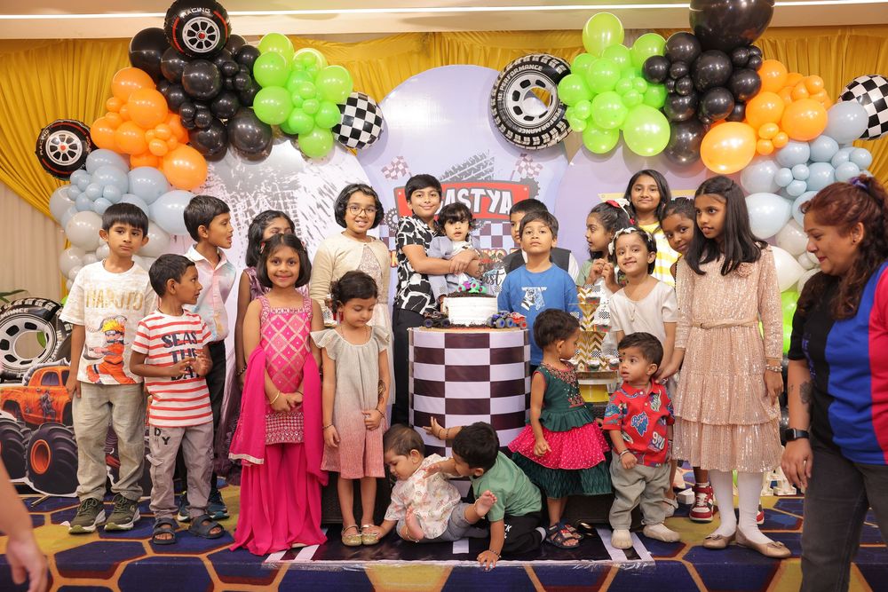 Children gather around a birthday cake at a colorful race car themed party with balloon decorations in orange, green, and black.
