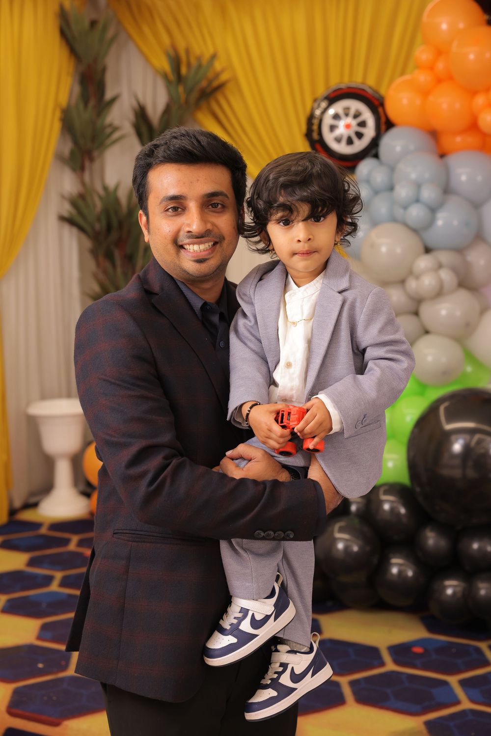 A man in a suit holds a young boy in a gray blazer at a colorful balloon-decorated birthday party.