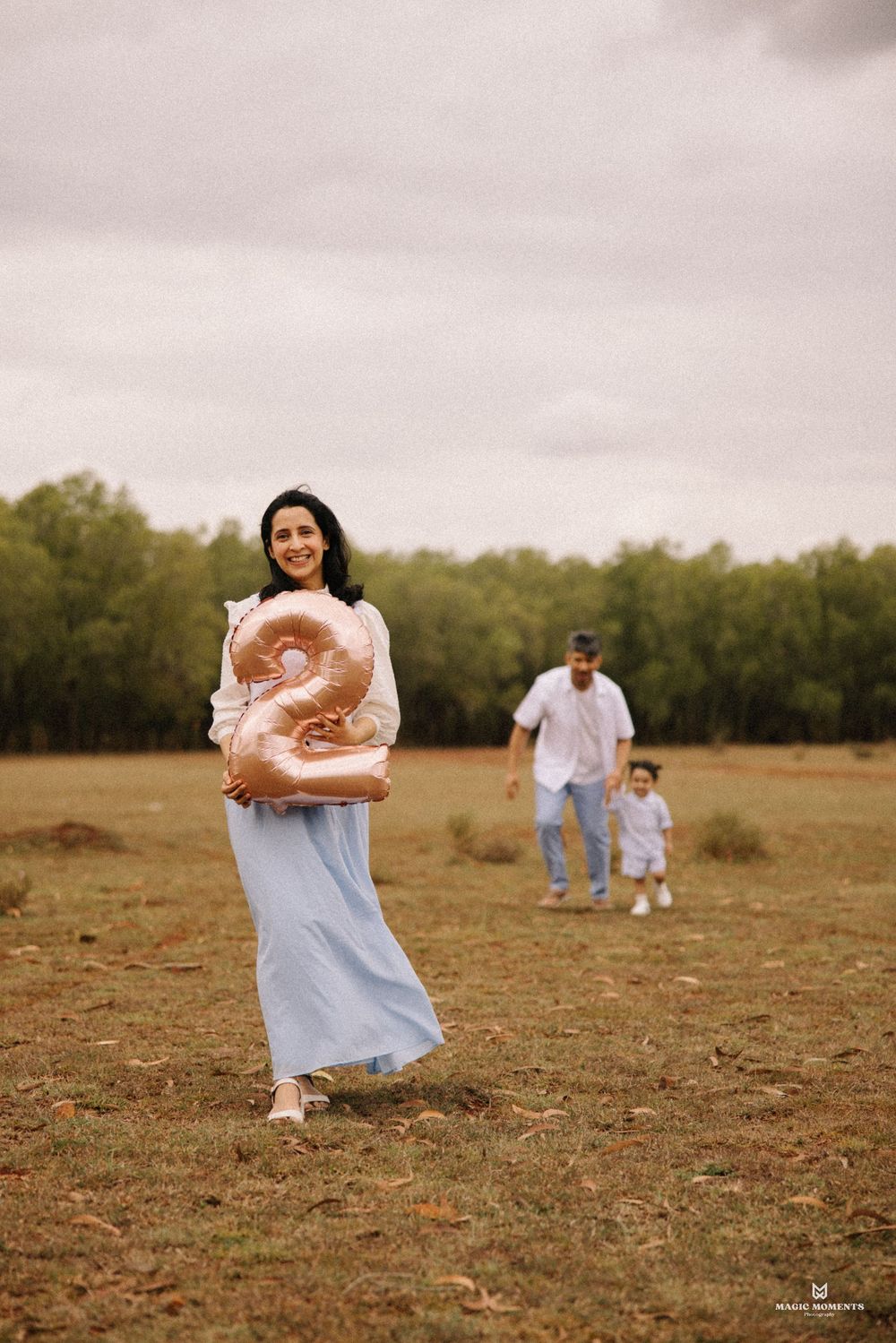 Woman holding a rose gold number 2 balloon in a field, with a man and toddler walking behind her.