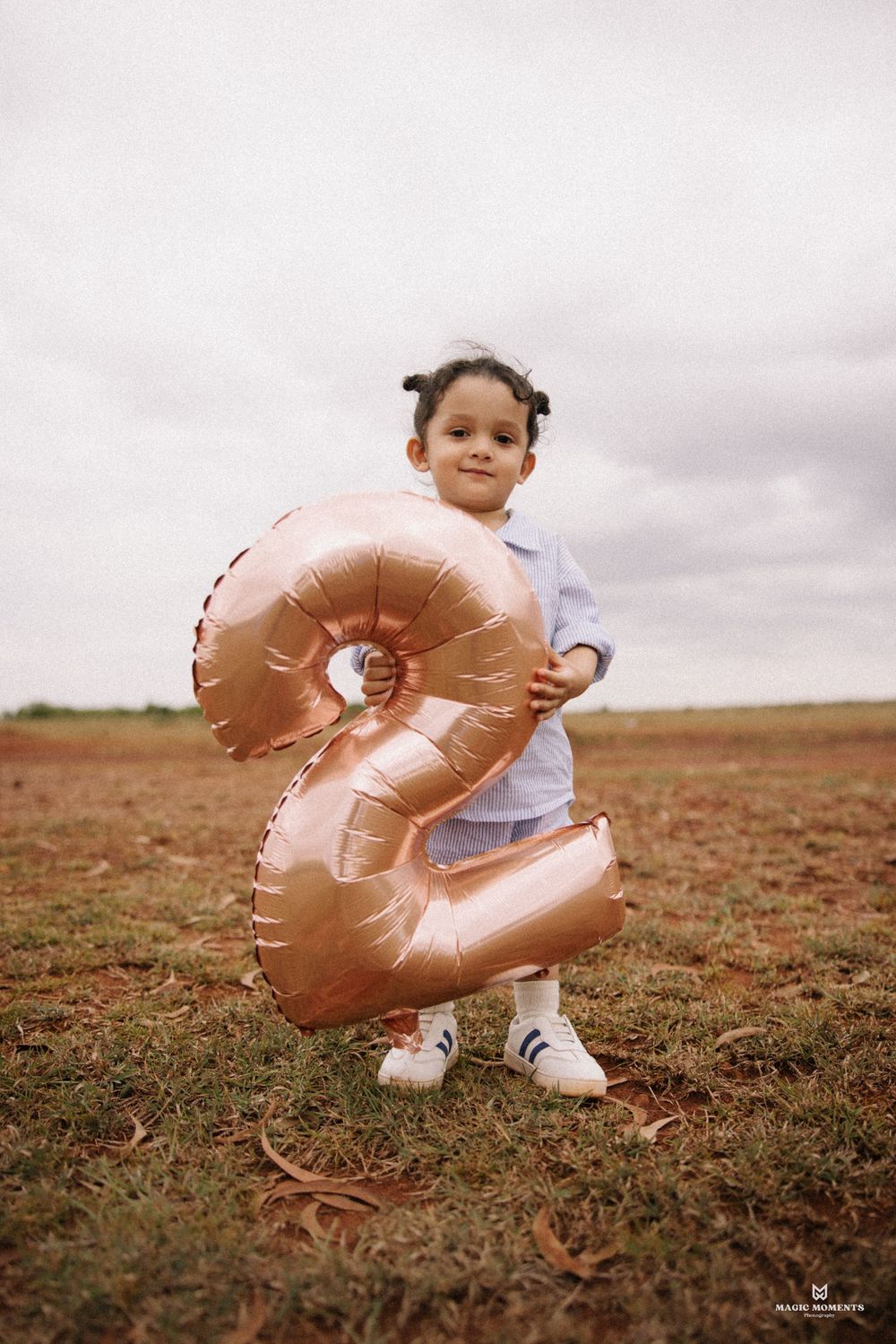 Toddler girl holding a rose gold number 2 balloon in a field, celebrating her 2nd birthday outdoors.