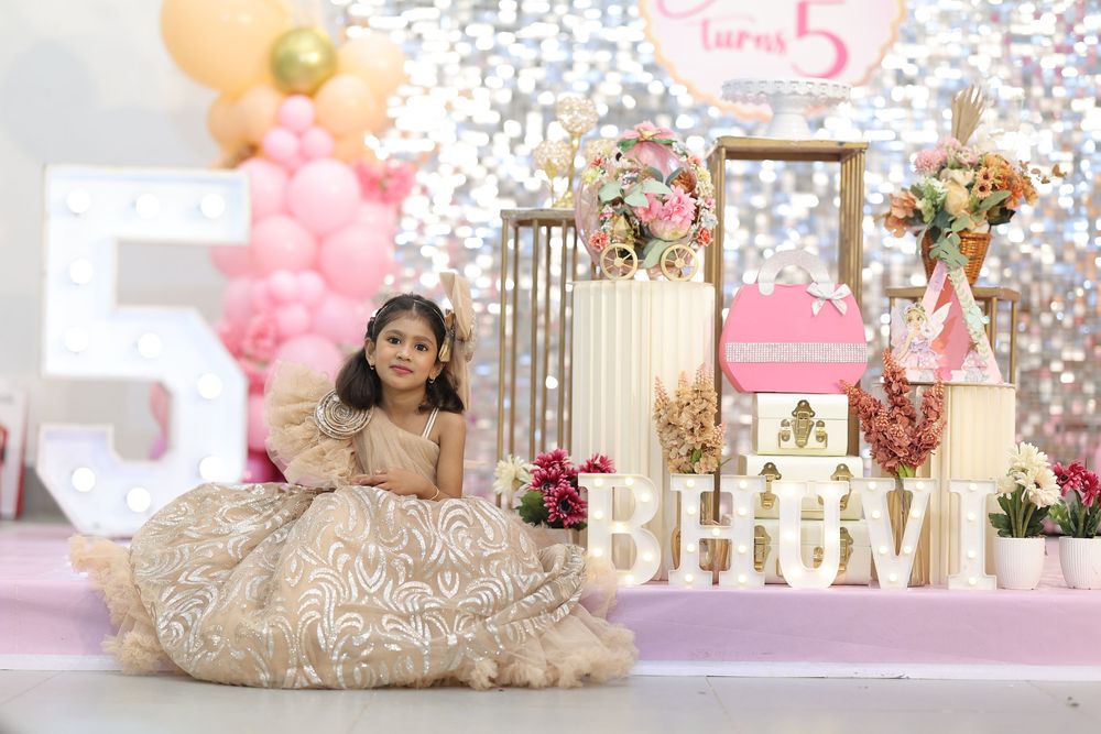 A young girl in a golden ballgown poses at her 5th birthday party with pink balloon decor and a decorative castle backdrop.