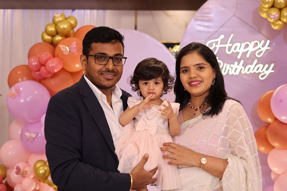 Family celebrating baby girl's first birthday party with pink and gold balloon decorations and Happy Birthday sign.