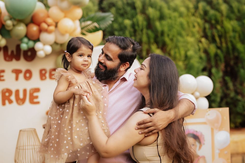 Family of three posing at a birthday party with balloon garland decorations in green and neutral tones.