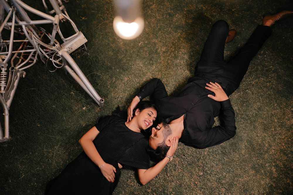 Couple in matching black outfits lying on grass, gazing at each other under a glowing bulb.