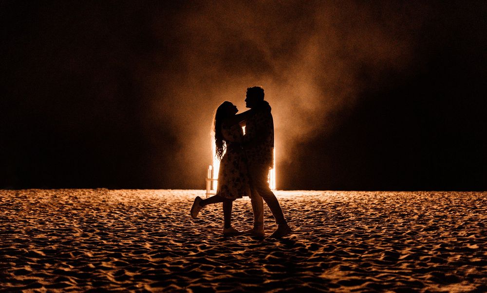 Romantic couple silhouette dancing on a beach at night with glowing fire light behind them.