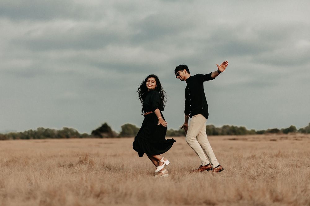Joyful couple holding hands and jumping in a golden open field under a cloudy sky.