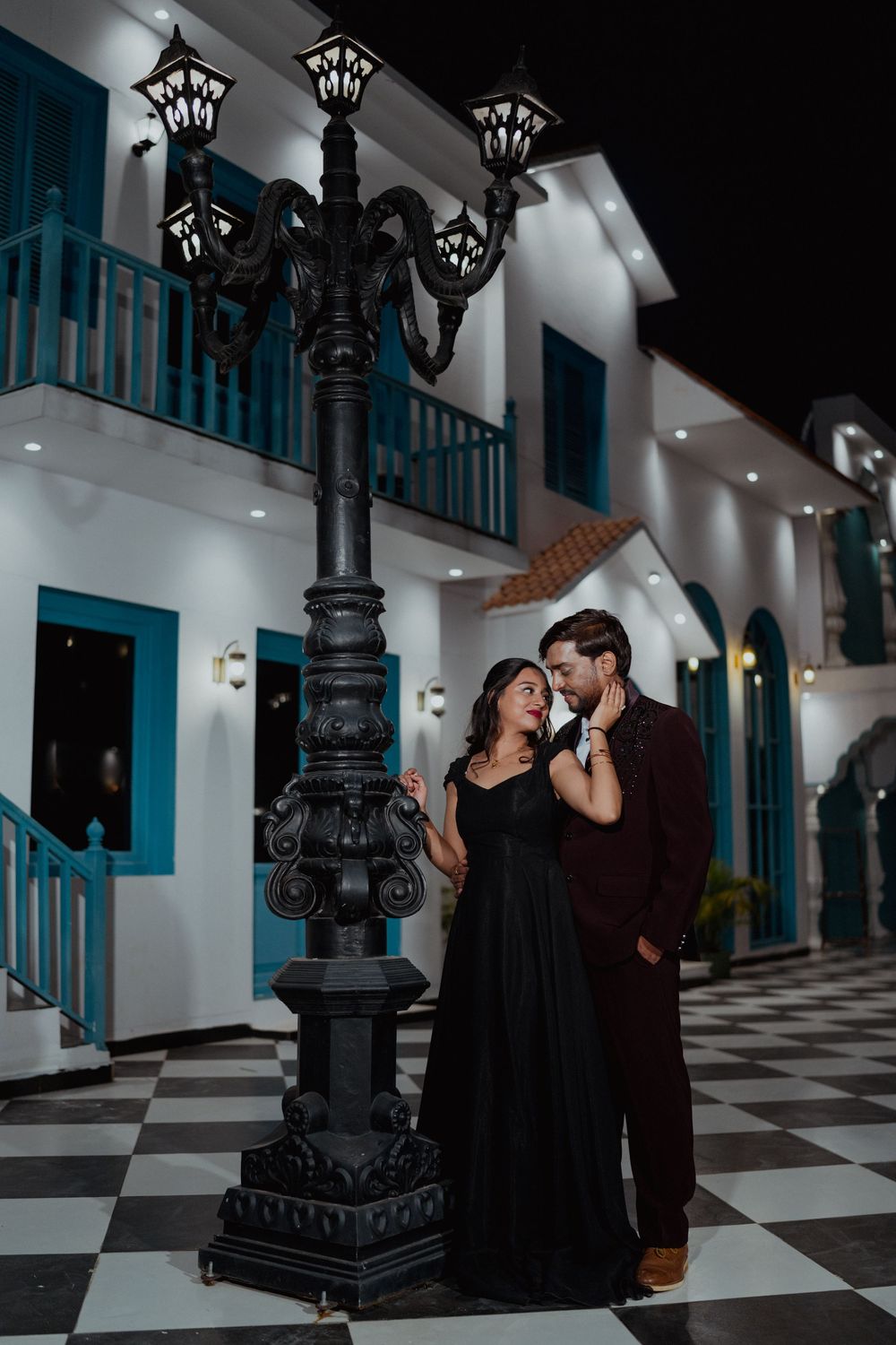 Couple in elegant attire posing by ornate street lamp at night outside white colonial building.