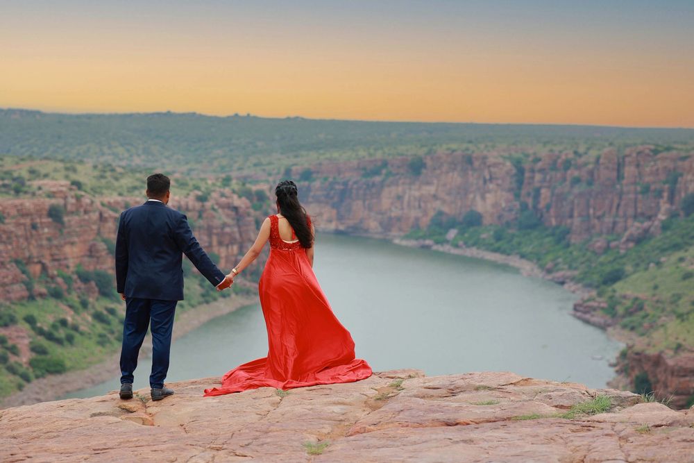 Couple holding hands on a cliff edge overlooking a scenic river canyon at sunset, woman in red gown.