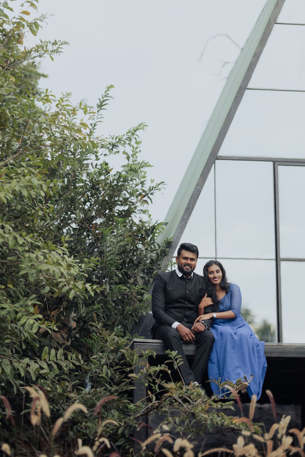 Couple posing outdoors near modern glass building surrounded by lush green trees, woman in blue dress, man in black suit.