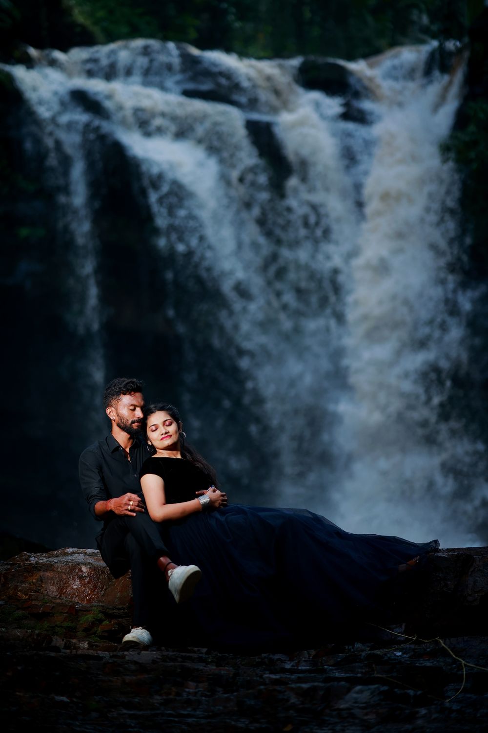 Couple sitting together in front of a powerful waterfall, both dressed in elegant black outfits.